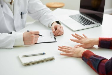 Hastanedeki bir randevu sırasında doktorun yanında el kol hareketi yapan bir hastanın parmaklarıyla çekilmiş bir fotoğrafı. Bir defter, bir dosya, bir dizüstü bilgisayar doktorun ofisindeki bir masada yatıyor.