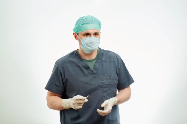 Ameliyat odasında ameliyat önlüğü giymiş bir cerrah elinde neşterle bekliyor. Cerrahi gömlek giyen, tek kullanımlık eldiven takan, tıbbi maske takan ve kep takan bir doktorun arkasında beyaz bir geçmiş..