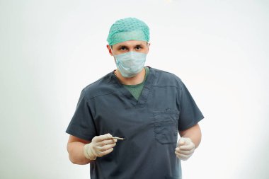 Ameliyat odasında ameliyat önlüklü bir cerrah neşterle poz veriyor. Cerrahi gömlek giyen, tek kullanımlık eldiven takan, tıbbi maske takan ve kep takan bir doktorun arkasında beyaz bir geçmiş..