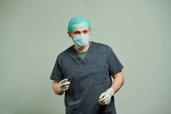 A male surgeon in scrubs is preparing a scalpel to do an incision of a patient in an operating room. A doctor wears a V neck surgical shirt, disposable medical gloves, a medical face mask, and a cap.