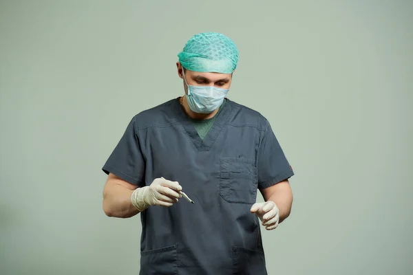 A surgeon in scrubs is checking to do an incision of a patient with a scalpel in an operating room. A doctor wears a surgical shirt, disposable medical gloves, a medical face mask, and a scrub cap.