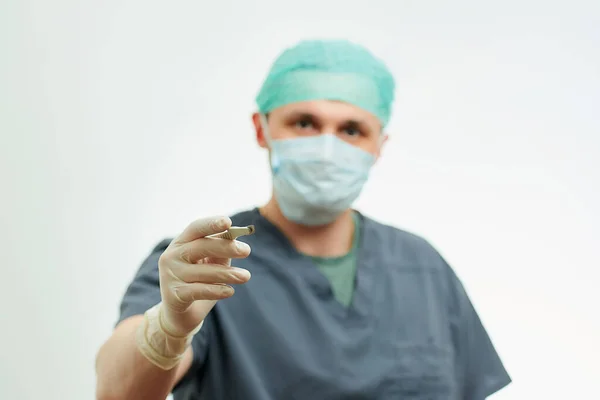A photo of a surgeon in scrubs with a focus on a scalpel in an operating room. White background behind a doctor who wears a surgical shirt, disposable gloves, a medical face mask, and a scrub cap.