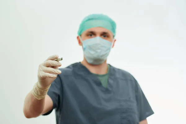 A photo of a surgeon in scrubs with a focus on a scalpel in a hospital. White background behind a doctor who wears a surgical shirt, disposable gloves, a medical face mask, and a scrub cap.