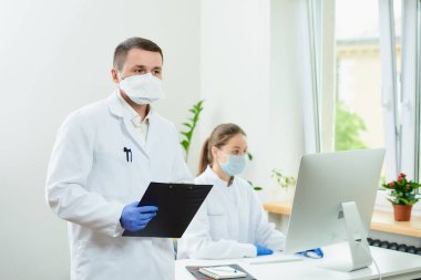 Ciddi bir cerrah tek kullanımlık eldiven ve solunum cihazı takar. Elinde hastane evrakları olan siyah bir dosya tutar. Kadın bir doktor köşedeki masaüstü bilgisayarının önünde oturuyor..  