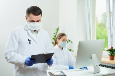 Tüylü bir cerrah solunum cihazı takar siyah bir pano tutar ve hastanede hastanın kartını doldurur. Kadın bir doktor odanın köşesindeki masaüstü bilgisayarının önünde oturuyor. 