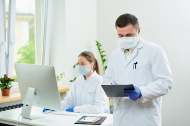 Bir cerrah solunum cihazı takar ve hastanede tek kullanımlık tıbbi eldiven hastanın kartını doldurur. Kadın bir doktor odanın köşesindeki masaüstü bilgisayarının önünde oturuyor.. 