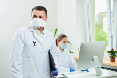 Tüylü bir cerrah solunum cihazı takar siyah bir dosya tutar ve hastaları hastanede bekler. Kadın bir doktor odanın köşesindeki masaüstü bilgisayarının önünde oturuyor..