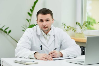 Beyaz laboratuvar önlüklü beyaz bir doktor hasta kartına teşhis koyup hastaneyi dinliyor. Laptopun yanında bir terapist doktor muayenehanesinde hasta bekliyor.. 