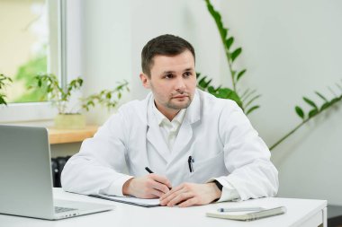 Beyaz laboratuvar önlüklü beyaz bir doktor bir hasta kartını teşhis koyarak hastanede düşünüyor. Laptopun yanında bir terapist doktor muayenehanesinde hasta bekliyor..