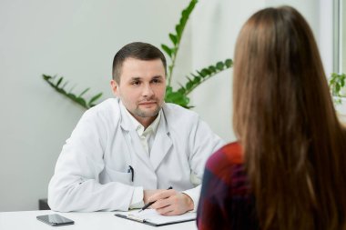 Beyaz laboratuvar önlüklü beyaz bir doktor bir masada oturuyor ve bir doktorun ofisinde kadın bir hastanın şikayetlerini yazıyor. Hastanede doktor randevusunda uzun saçlı bir kadın..