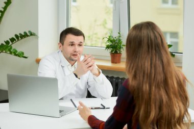 Beyaz laboratuvar önlüklü beyaz bir doktor bir masada oturuyor ve bir doktorun ofisinde bir kadın hastayla tedaviyi tartışıyor. Hastanede doktor randevusunda uzun saçlı bir kadın..