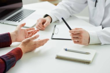Kadın bir hastanın ve bir doktorun hastanedeki randevularında çekilmiş yakın bir fotoğrafı. İçinde kalem, dosya, dizüstü bilgisayar olan bir defter doktorun ofisinde beyaz bir masada yatıyor..