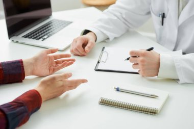 Hastanedeki randevu sırasında bir kadın hastayla bir doktorun ellerinin yakın bir fotoğrafı. İçinde kalem, dosya, dizüstü bilgisayar olan bir defter doktorun ofisinde beyaz bir masada yatıyor..