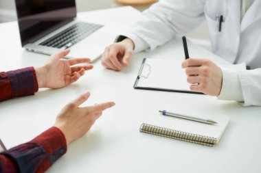 Kadın bir hastayla bir doktorun yakın bir fotoğrafı hastanede yapılan bir randevu sırasında el kol hareketi yapıyor. İçinde kalem, dosya, dizüstü bilgisayar olan bir defter doktorun ofisindeki bir masada yatıyor..