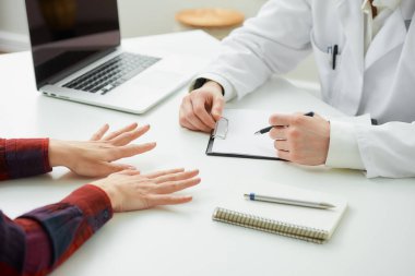Hastanedeki bir randevu sırasında doktorun yanında el kol hareketi yapan bir hastanın parmaklarıyla çekilmiş bir fotoğrafı. Bir defter, bir dosya, bir dizüstü bilgisayar doktorun ofisindeki bir masada yatıyor.