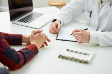 Hastanedeki bir randevu sırasında doktorun yanında el kol hareketi yapan bir hastanın katlanmış ellerinin fotoğrafı. İçinde kalem, dosya, dizüstü bilgisayar olan bir defter doktorun ofisinde beyaz bir masada yatıyor.