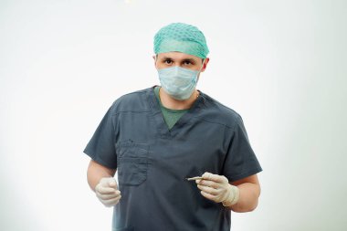 Ameliyat odasında ameliyat önlüklü bir cerrah neşterle poz veriyor. Cerrahi gömlek giyen, tek kullanımlık eldiven takan, tıbbi maske takan ve kep takan bir doktorun arkasında beyaz bir geçmiş..