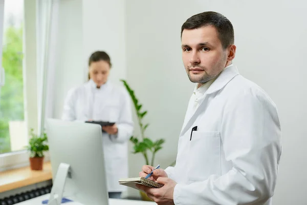 A serious caucasian doctor with a notebook and a pen records the patients medical history in a hospital. A female therapist is doing notes in a clipboard near an all in one computer in a clinic.