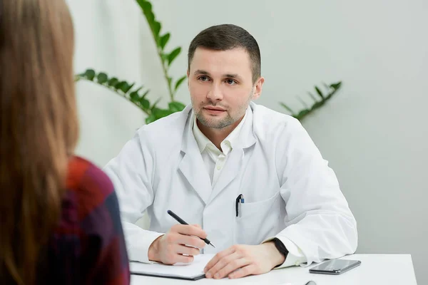 A portrait of a doctor in a white lab coat who is sitting at a desk and listening to a female patient in a doctor\'s office. A woman at a doctor\'s appointment in a hospital.