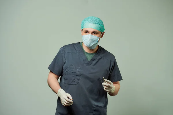 A male surgeon in scrubs is preparing a scalpel to do an incision of a patient in an operating room. A doctor wears a V neck surgical shirt, disposable medical gloves, a medical face mask, and a cap.