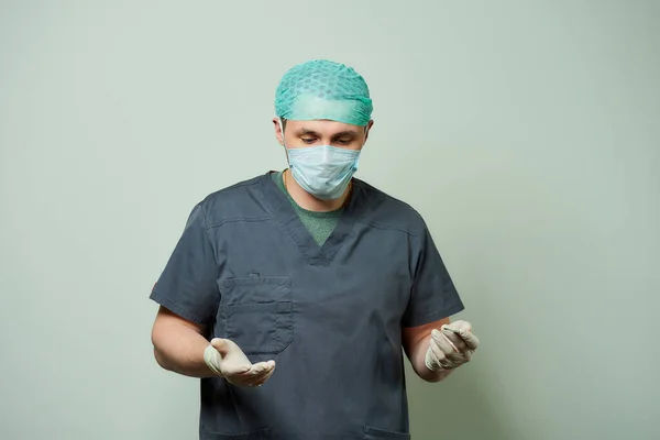 A caucasian male surgeon is holding a scalpel in a hospital. A doctor wears a V neck surgical shirt, disposable medical gloves, a medical face mask, and a scrub cap in an operating room.