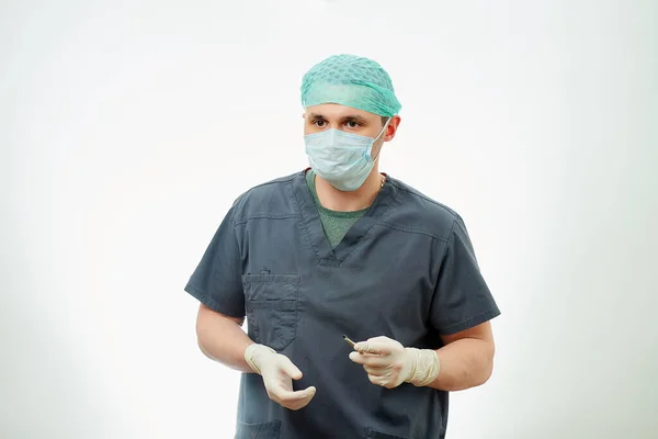 A surgeon in scrubs is standing with a scalpel in an operating room. White background behind a doctor who wears a surgical shirt, disposable medical gloves, a medical face mask, and a scrub cap.