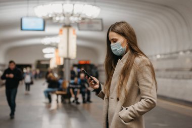 Koronavirüsün yayılmasını önlemek için tıbbi maske takan bir kadın metro istasyonunda akıllı telefon kullanıyor. Yüzünde ameliyat maskesi olan bir kız COVID-19 'a karşı cep telefonundan haber okuyor..