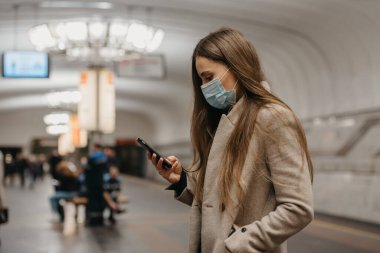 Koronavirüsün yayılmasını önlemek için tıbbi maske takan bir kadın metro istasyonunda akıllı telefon kullanıyor. Yüzünde cerrahi maske olan bir kız COVID-19 'a karşı cep telefonunda haberlere çıkıyor.