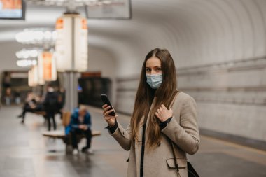 Coronavirus 'un yayılmasını önlemek için maskeli bir kadın metro istasyonunda akıllı telefonuyla poz veriyor. COVID-19 'a karşı cerrahi maske takan bir kız metro istasyonunda tren bekliyor..