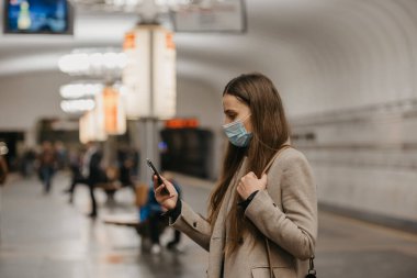 Koronavirüsün yayılmasını önlemek için maske takan bir kadın metro istasyonunda akıllı telefon kullanıyor. COVID-19 'a karşı cerrahi maske takan bir kız metro istasyonunda tren bekliyor..