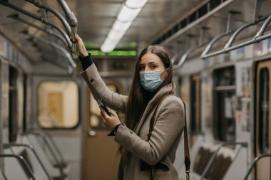 Koronavirüsün yayılmasını önlemek için tıbbi maske takan bir kadın metro vagonunda akıllı telefon tutuyor. COVID-19 'a karşı ameliyat maskesi takan bir kız trende cep telefonuyla haberlere çıkıyor..