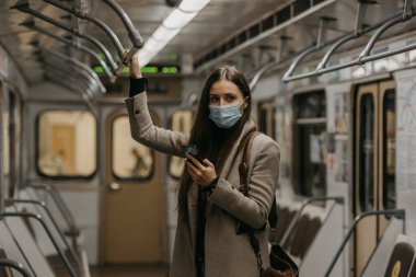 Koronavirüsün yayılmasını önlemek için tıbbi maske takan bir kadın metro vagonunda poz veriyor. Yüzünde ameliyat maskesi olan uzun saçlı bir kız COVID-19 'a karşı bir trende cep telefonu tutuyor..