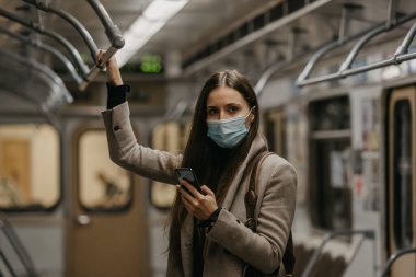 Koronavirüsün yayılmasını önlemek için tıbbi maske takan bir kadın metro vagonunda poz veriyor. Yüzünde COVID-19 maskesi olan bir kız trende cep telefonu tutuyor..