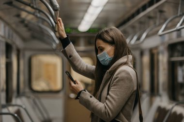 Koronavirüsün yayılmasını önlemek için tıbbi maske takan bir kadın metro vagonunda akıllı telefon kullanıyor. COVID-19 'a karşı ameliyat maskesi takan bir kız trende cep telefonuyla haberlere çıkıyor..