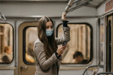 Coronavirus 'un yayılmasını önlemek için maskeli bir kadın metro vagonunda telefon kullanıyor. Yüzünde ameliyat maskesi olan uzun saçlı bir kız COVID-19 'a karşı bir trende cep telefonu tutuyor..