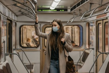 Coronavirus 'un yayılmasını önlemek için maskeli bir kadın metro vagonunun ortasındaki tırabzana tutunuyor. COVID-19 'a karşı cerrahi maskeli güzel bir kız bir metro treninde duruyor..
