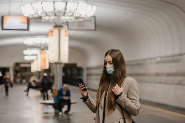 A woman in a face mask to avoid the spread of coronavirus is reading news on a phone at a subway station. A girl in a surgical face mask against COVID-19 is waiting for a train on a metro platform.