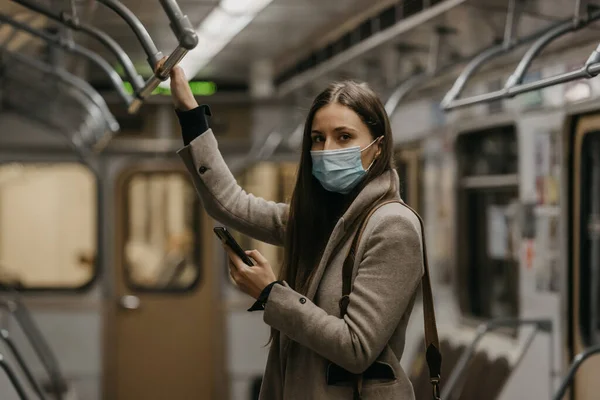 A woman in a medical face mask to avoid the spread of coronavirus is looking around in a subway car. A girl in a surgical mask on her face against COVID-19 is holding a cellphone on a train.