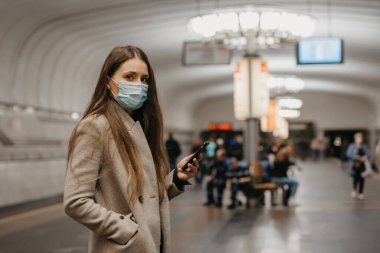 Coronavirus 'un yayılmasını önlemek için tıbbi maske takan bir kadın metro istasyonunda akıllı telefon tutuyor. Yüzünde ameliyat maskesi olan bir kız COVID-19 'a karşı cep telefonunu kullanıyor..