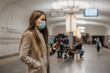 Koronavirüsün yayılmasını önlemek için maskeli bir kadın metro istasyonunda akıllı telefon kullanıyor. Yüzünde cerrahi maske olan bir kız COVID-19 'a karşı metro istasyonunda tren bekliyor.