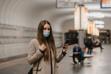 Coronavirus 'un yayılmasını önlemek için maskeli bir kadın metro istasyonunda akıllı telefonuyla poz veriyor. COVID-19 'a karşı cerrahi maske takan bir kız metro istasyonunda tren bekliyor..