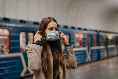 Bir kadın metroda koronavirüsün yayılmasını önlemek için yüzüne tıbbi maske takıyor. Uzun saçlı, bej ceketli bir kız metro istasyonunda tren bekliyor..