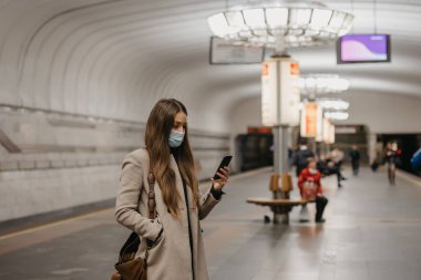 Coronavirus 'un yayılmasını önlemek için maske takan bir kadın metro istasyonunda akıllı telefon tutuyor. Yüzünde cerrahi maske olan bir kız COVID-19 'a karşı metro istasyonunda tren bekliyor.