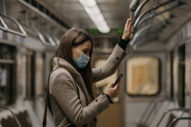 Koronavirüsün yayılmasını önlemek için tıbbi maske takan bir kadın metro vagonunda akıllı telefon kullanıyor. COVID-19 'a karşı ameliyat maskesi takan bir kız trende cep telefonuyla haberlere çıkıyor..