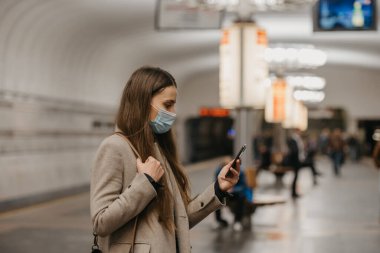Koronavirüsün yayılmasını önlemek için maske takan bir kadın metro istasyonunda akıllı telefon kullanıyor. COVID-19 'a karşı cerrahi maske takan bir kız metro istasyonunda tren bekliyor..