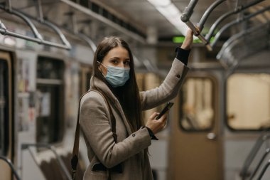 Coronavirus 'un yayılmasını önlemek için tıbbi maske takan bir kadın metro vagonuna bakıyor. Yüzünde COVID-19 maskesi olan bir kız trende cep telefonu tutuyor..