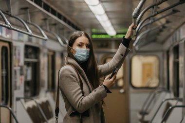 Koronavirüsün yayılmasını önlemek için tıbbi maske takan bir kadın metro vagonunda akıllı telefon tutuyor. COVID-19 'a karşı ameliyat maskesi takan bir kız trende cep telefonuyla haberlere çıkıyor..