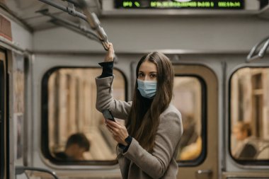 Coronavirus 'un yayılmasını önlemek için maske takan bir kadın bir metro vagonuna bakıyor. Yüzünde ameliyat maskesi olan uzun saçlı bir kız COVID-19 'a karşı bir trende cep telefonu tutuyor..