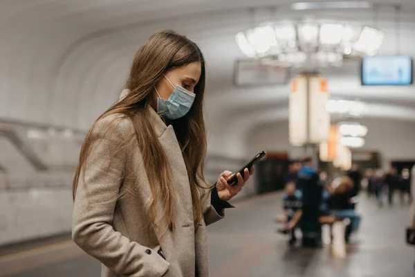 A woman in a medical face mask to avoid the spread of coronavirus is using a smartphone at the subway station. A girl in a surgical mask on the face against COVID-19 is scrolling news on her cellphone