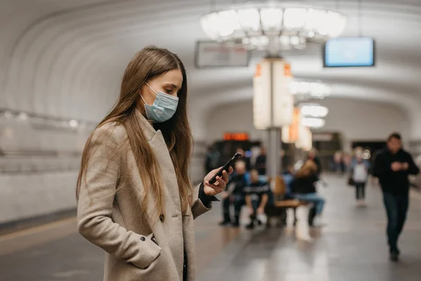 A woman in a medical face mask to avoid the spread of coronavirus is using a smartphone at the subway station. A girl in a surgical mask on the face against COVID-19 is reading news on her cellphone.